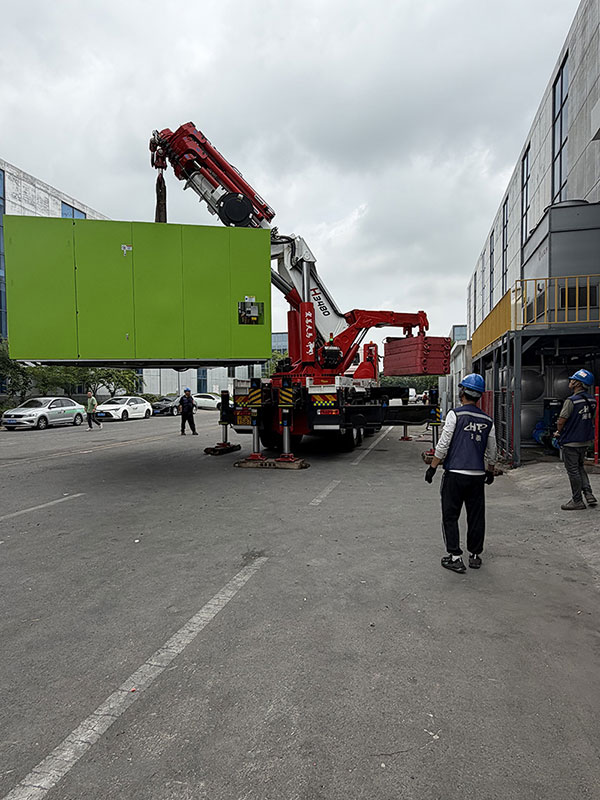 两栋厂房之间的厂区道路上折臂吊起吊绿色大型设备，多名作业人员协同指挥